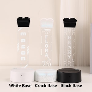 May include: Three personalized LED night lights with different base colors: white, crack, and black. Each light features a unique name etched into a clear acrylic panel, topped with a black decorative element. The bases are labeled "White Base", "Crack Base", and "Black Base".