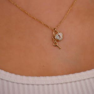 Puede incluir: Un collar dorado con un colgante en forma de corazón con diseño de cerradura, adornado con pequeñas piedras brillantes. Un pequeño dije de llave cuelga debajo. El collar se muestra sobre un fondo neutro.