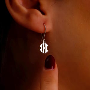May include: A silver-colored monogram earring with a circular design, hanging from a hook. The earring features the intertwined initials "IHC" in a stylized font. The earring is shown on a person's ear.
