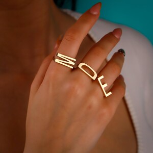 May include: Gold-colored rings spelling out the word "MODE" are displayed on a hand. The rings are minimalist in design, with each letter crafted in a simple, blocky font. The rings are worn on different fingers.