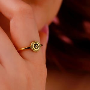 May include: A gold ring with a circular black emblem featuring the letters "AES" in a serif font. The ring has a thin band and a decorative border around the emblem. The ring is on a finger.
