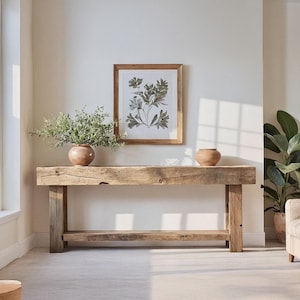 May include: A rustic wooden console table with a thick rectangular top and sturdy legs, positioned against a white wall. On the table sit two terracotta-colored ceramic vases, one containing green foliage, beneath a framed botanical print.