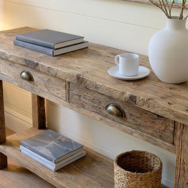 May include: A rustic wooden console table with two drawers and brass hardware. The table is topped with books, a white teacup and saucer, and a white vase. A woven basket sits below.