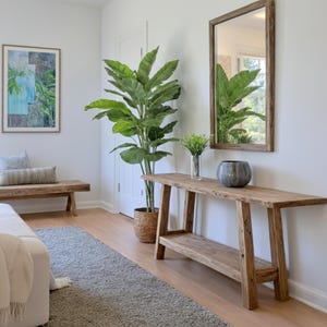 Può includere: Una consolle in legno rustico con un ripiano inferiore e gambe angolate. Una grande pianta in vaso con foglie verdi è accanto al tavolo. Uno specchio incorniciato e un'opera d'arte sono appesi alla parete bianca. Un tappeto grigio e una panca completano l'arredamento della stanza.