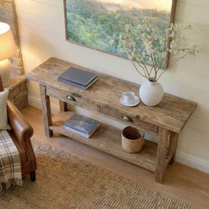May include: A rustic wooden console table with two drawers and a lower shelf. The table is adorned with books, a white vase with branches, a small woven basket, and a teacup. A framed landscape painting hangs above.