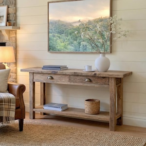 Peut inclure: Une console en bois rustique avec deux tiroirs et une étagère inférieure. La table est en bois clair et a un aspect naturel et vieilli. Un panier en osier, des livres et un vase blanc avec des fleurs sont sur la table.