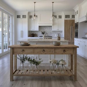 Peut inclure: Îlot de cuisine en bois clair avec trois tiroirs et une étagère inférieure, contenant des bouteilles en verre avec de la verdure et des fleurs. La cuisine est lumineuse, blanche, avec un plan de travail en marbre et des suspensions.
