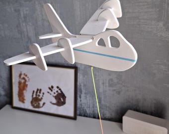 May include: A white wooden airplane mobile with blue accents and a colorful string. The toy plane hangs from the ceiling. A framed picture with handprints and footprints is on a white surface.
