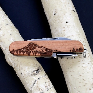 Puede incluir: Un cuchillo de bolsillo de madera con un diseño grabado con láser de una cordillera. El cuchillo tiene una hoja plateada y un mango de madera marrón.