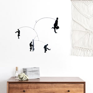 Könnte beinhalten: Eine moderne Mobile-Skulptur mit schwarzen Silhouette-Figuren in verschiedenen Posen, die an dünnen Drähten hängen. Die Figuren scheinen Geschäftsleute zu sein, einige tragen Aktentaschen. Die Skulptur ist an einer weißen Wand angebracht, mit einer Holzkommode und dekorativen Gegenständen im Vordergrund.