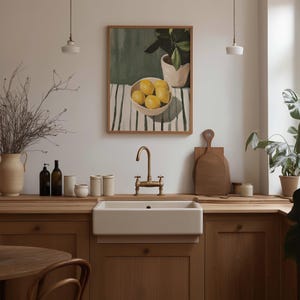 May include: A kitchen scene featuring a farmhouse sink with a brass faucet, wooden countertops, and cabinets. A framed painting of lemons in a bowl hangs above the sink. Decorative items include vases, bottles, and cutting boards.