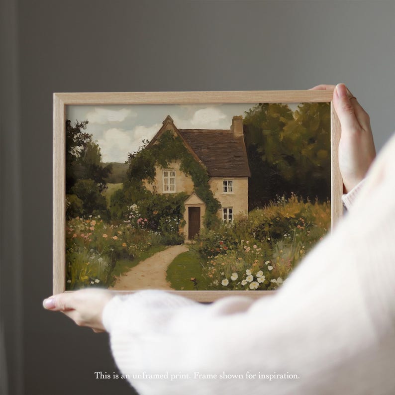 May include: A framed print of a cottage scene. The artwork depicts a stone cottage with a thatched roof, covered in ivy, and surrounded by a garden path and colorful flowers. The frame is light-colored wood.