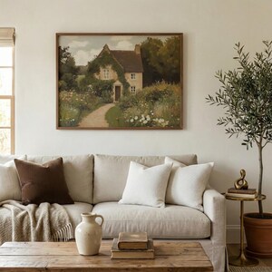 May include: A framed landscape painting of a cottage with a path leading to the front door. The painting hangs above a beige sofa with decorative pillows and a wooden coffee table. A small olive tree is to the right.