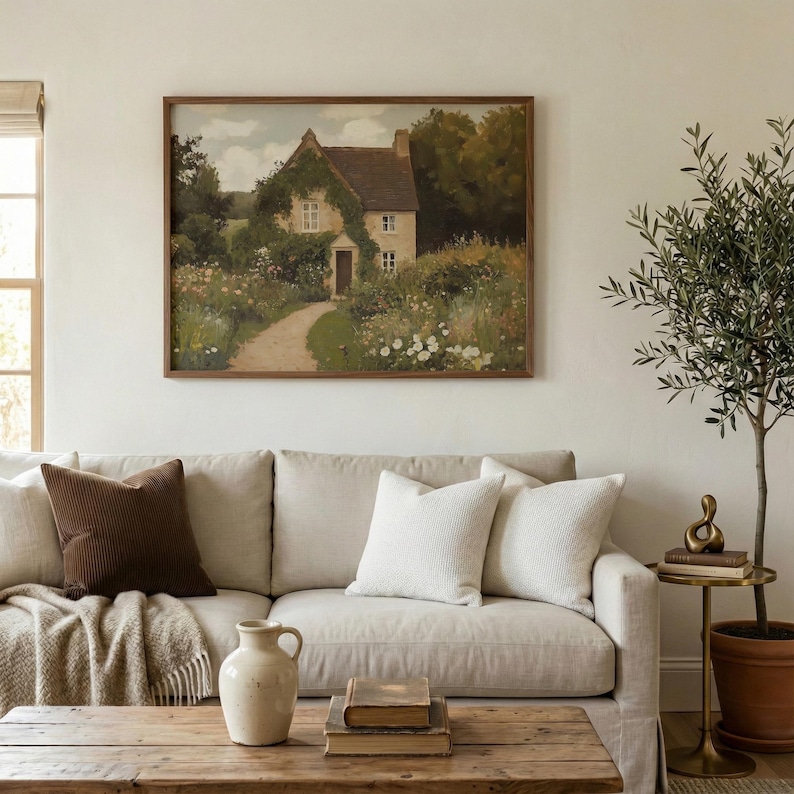 May include: A framed landscape painting of a cottage with a path leading to the front door. The painting hangs above a beige sofa with decorative pillows and a wooden coffee table. A small olive tree is to the right.