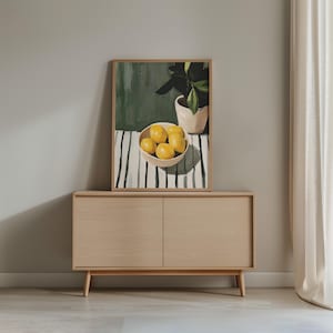 May include: A framed art print depicts a still life with lemons in a bowl, a potted plant, and a striped tablecloth. The artwork is displayed on a light wood cabinet with two doors. The background is a neutral gray wall.