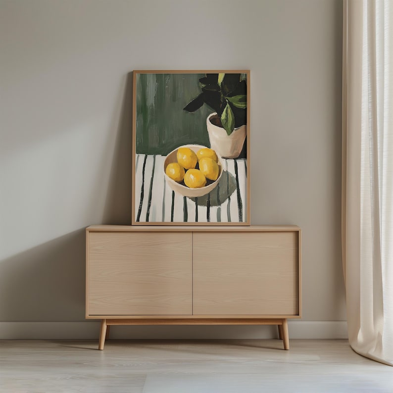 May include: A framed art print depicts a still life with lemons in a bowl, a potted plant, and a striped tablecloth. The artwork is displayed on a light wood cabinet with two doors. The background is a neutral gray wall.