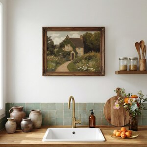 May include: A kitchen scene featuring a framed landscape painting of a cottage with a path. A gold faucet and white sink are in the foreground. Decorative jars, wooden cutting boards, and a vase of flowers add to the rustic aesthetic.