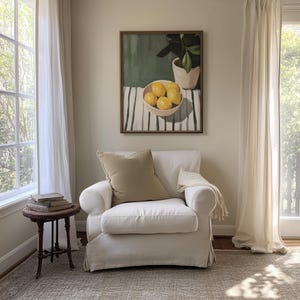 May include: A cozy living room scene featuring a white armchair with a tan pillow and a small, dark wooden side table. A framed painting of lemons in a bowl hangs above the chair. Sheer white curtains frame a large window, creating a bright and airy atmosphere.