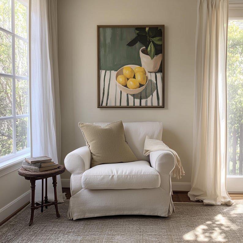 May include: A cozy living room scene featuring a white armchair with a tan pillow and a small, dark wooden side table. A framed painting of lemons in a bowl hangs above the chair. Sheer white curtains frame a large window, creating a bright and airy atmosphere.