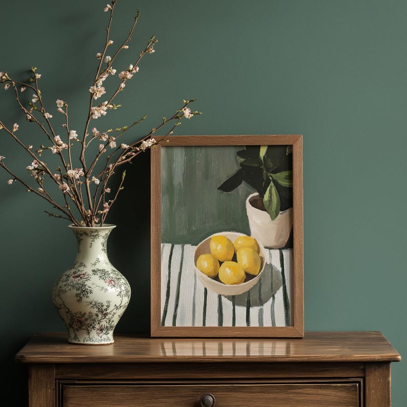 May include: A still life painting in a wooden frame depicts a bowl of lemons, a potted plant, and a striped tablecloth. A floral vase with flowering branches sits to the left. The scene is set on a wooden surface against a dark green wall.