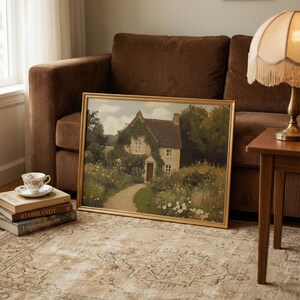 May include: A framed landscape painting of a cottage with a path leading to the front door. The painting has a gold frame and is propped against a brown corduroy sofa. A teacup and books are in the foreground.