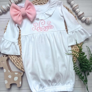 May include: A white long-sleeve infant gown featuring a ruffled neckline, bell sleeves, and a pink embroidered monogram on the chest. A large, textured pink fabric bow is attached to the shoulder, complementing the embroidery.