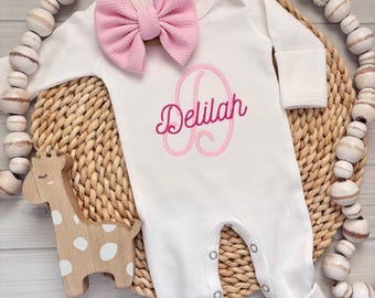May include: White baby onesie with the name "Delilah" embroidered in pink, along with a pink bow. A wooden giraffe toy and a beaded garland are also in the image. The onesie has long sleeves and snaps.
