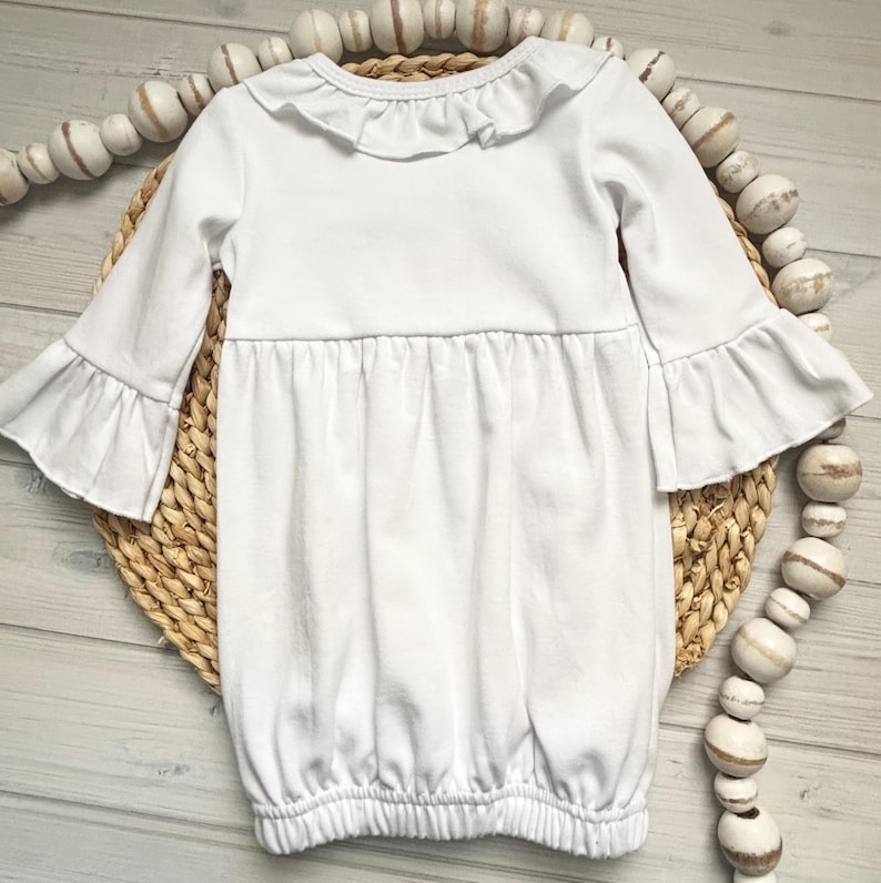 May include: A solid white long-sleeve gown featuring a ruffled collar and flared bell sleeves. The garment has a gathered waistline and an elasticized bottom hem, creating a soft, flowing silhouette with a simple, clean design.