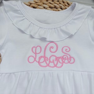 May include: A white long-sleeve shirt featuring a ruffled collar and a pink embroidered monogram on the chest. The garment has a gathered waistline and long sleeves with ruffled cuffs, showcasing a detailed script font for the lettering.