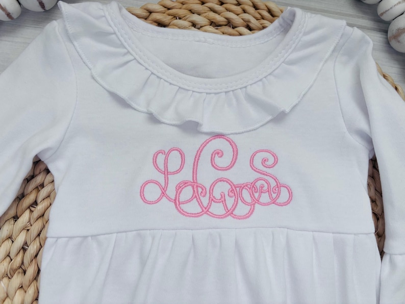 May include: A white long-sleeve shirt featuring a ruffled collar and a pink embroidered monogram on the chest. The garment has a gathered waistline and long sleeves with ruffled cuffs, showcasing a detailed script font for the lettering.