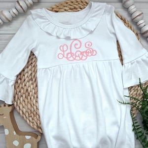 May include: A white long-sleeved gown featuring a ruffled collar and ruffled sleeve cuffs. The center of the chest displays a pink embroidered monogram with cursive lettering. The garment has a gathered waistline and a loose, flowing skirt.