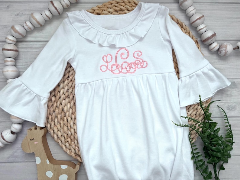 May include: A white long-sleeved gown featuring a ruffled collar and ruffled sleeve cuffs. The center of the chest displays a pink embroidered monogram with cursive lettering. The garment has a gathered waistline and a loose, flowing skirt.