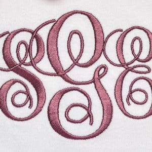 May include: A white fabric surface featuring a large, centered monogram embroidery in a dusty rose thread color. The design consists of three interconnected, stylized cursive letters with intricate loops and swirls, stitched in a satin stitch.