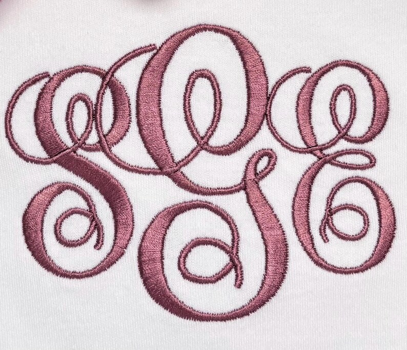 May include: A white fabric surface featuring a large, centered monogram embroidery in a dusty rose thread color. The design consists of three interconnected, stylized cursive letters with intricate loops and swirls, stitched in a satin stitch.