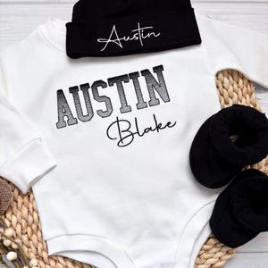 May include: A white baby onesie with the name "Austin Blake" embroidered in black. A matching black beanie with "Austin" embroidered in white is above the onesie. Two black baby booties are on the right side.