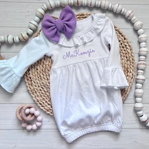 May include: A white long-sleeve gown with ruffled cuffs and a ruffled neckline. The gown features the name McKenzie embroidered in purple thread on the chest. A large purple textured bow is attached to the neckline of the white garment.