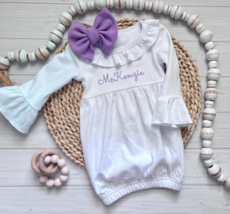 May include: A white long-sleeve gown with ruffled cuffs and a ruffled neckline. The gown features the name McKenzie embroidered in purple thread on the chest. A large purple textured bow is attached to the neckline of the white garment.