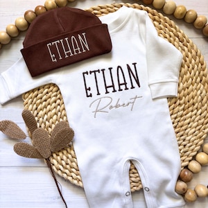 May include: A white baby onesie and matching brown beanie, both embroidered with the name "ETHAN". The onesie also has the name "Robert" embroidered below "ETHAN". The set is displayed on a woven mat with wooden beads.
