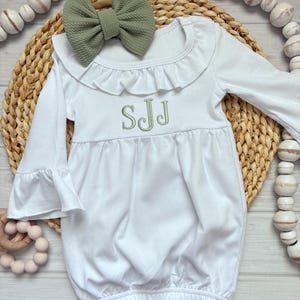 May include: A white long-sleeve gown with a ruffled collar and bell sleeves, featuring the embroidered green monogram SJJ on the chest. A matching sage green textured fabric bow is positioned at the neckline of the garment.