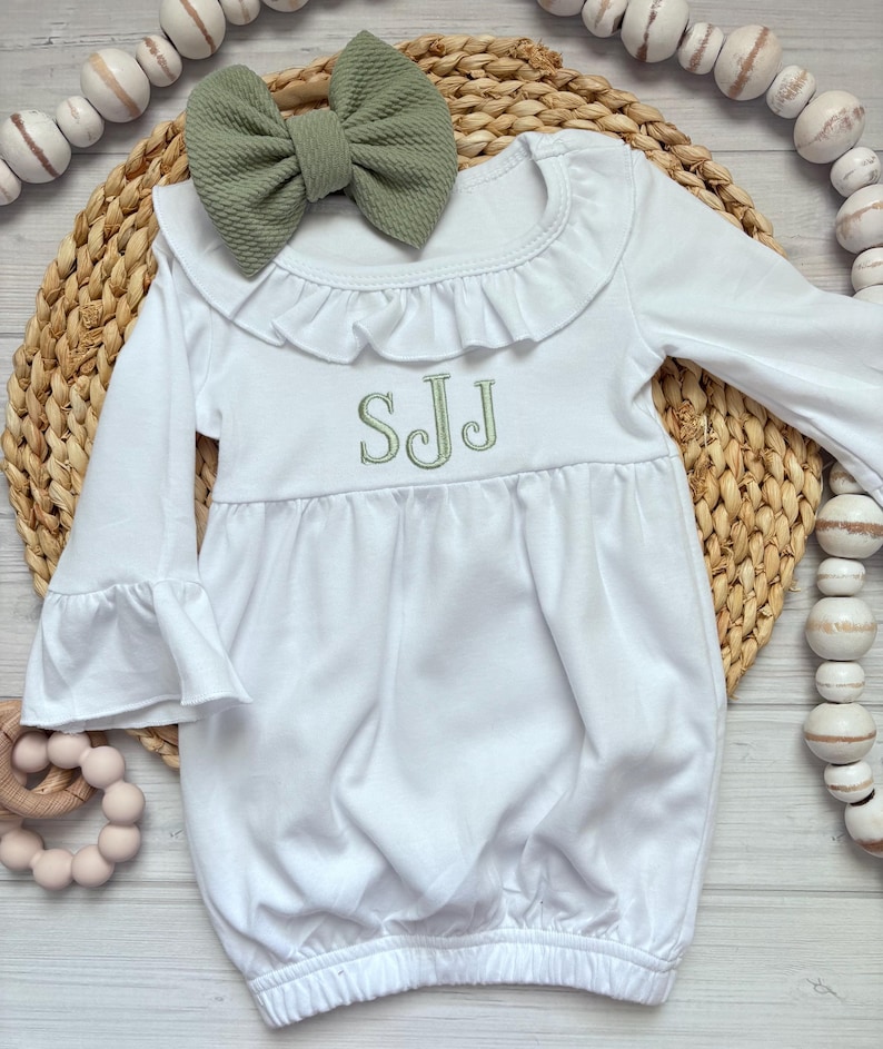 May include: A white long-sleeve gown with a ruffled collar and bell sleeves, featuring the embroidered green monogram SJJ on the chest. A matching sage green textured fabric bow is positioned at the neckline of the garment.