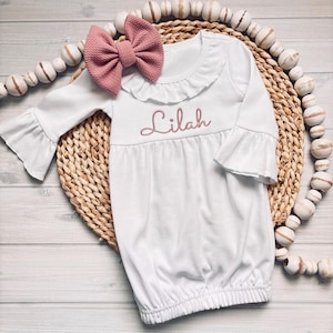 May include: A white long-sleeved baby gown featuring a ruffled neckline, bell sleeves, and the name Lilah embroidered in dusty rose thread across the chest. A matching dusty rose textured fabric bow is attached near the neckline.