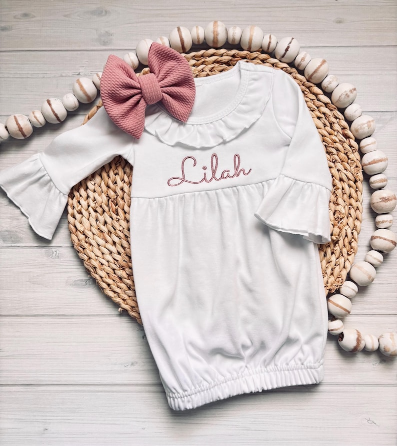 May include: A white long-sleeved baby gown featuring a ruffled neckline, bell sleeves, and the name Lilah embroidered in dusty rose thread across the chest. A matching dusty rose textured fabric bow is attached near the neckline.