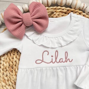 May include: A white long-sleeved dress with a ruffled collar and the name Lilah embroidered in dusty pink thread across the chest. A matching dusty pink textured fabric bow is positioned near the neckline of the garment.
