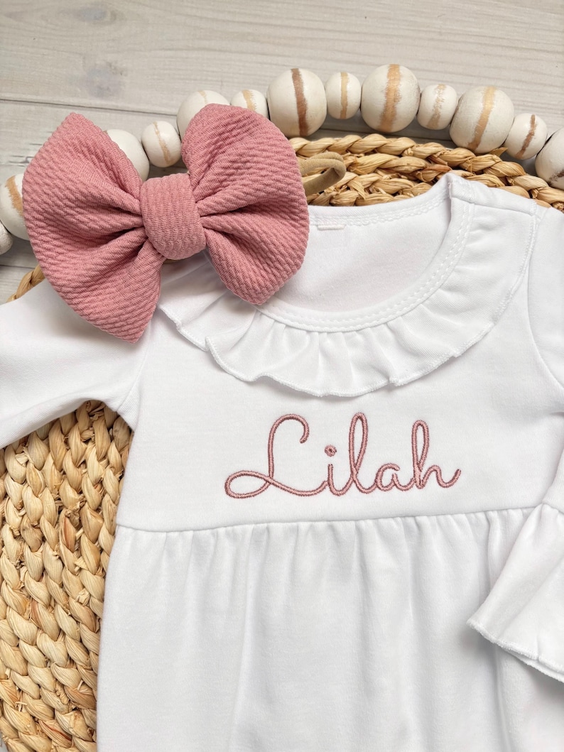 May include: A white long-sleeved dress with a ruffled collar and the name Lilah embroidered in dusty pink thread across the chest. A matching dusty pink textured fabric bow is positioned near the neckline of the garment.