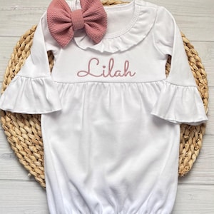 May include: A white long-sleeve gown with a ruffled neckline and bell sleeves. The name Lilah is embroidered in pink thread across the chest. A matching dusty pink textured fabric bow is attached to the neckline of the garment.