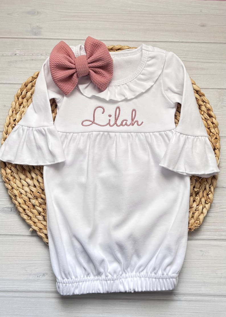 May include: A white long-sleeve gown with a ruffled neckline and bell sleeves. The name Lilah is embroidered in pink thread across the chest. A matching dusty pink textured fabric bow is attached to the neckline of the garment.