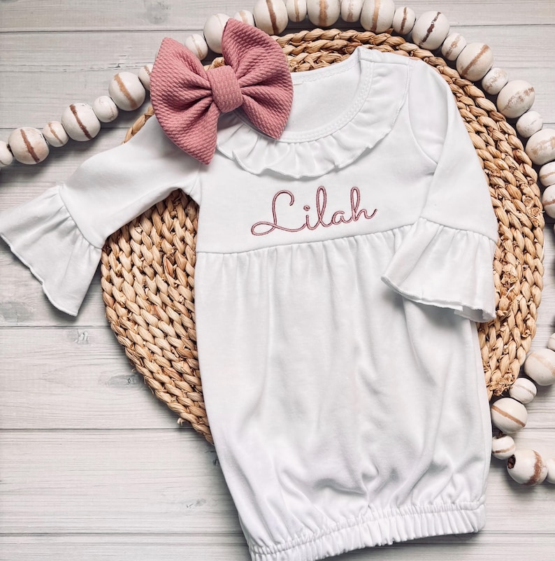 May include: A white long-sleeved gown with a ruffled collar and bell sleeves, featuring the name Lilah embroidered in dusty pink thread on the chest. A large, matching dusty pink textured fabric bow is attached to the neckline.