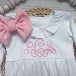 May include: A white long-sleeve dress featuring a ruffled neckline and a pink embroidered monogram on the chest. A large, textured pink fabric bow is attached to the shoulder. The garment has a gathered waist and long sleeves with ruffled cuffs.
