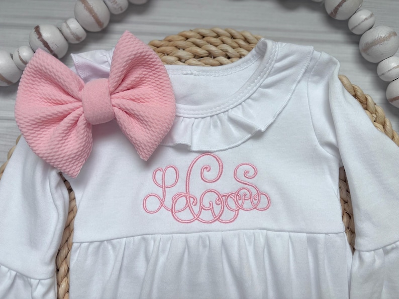 May include: A white long-sleeve dress featuring a ruffled neckline and a pink embroidered monogram on the chest. A large, textured pink fabric bow is attached to the shoulder. The garment has a gathered waist and long sleeves with ruffled cuffs.