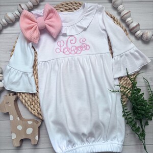 May include: A white long-sleeve infant gown featuring a ruffled collar, bell sleeves, and a pink embroidered monogram on the chest. The gown is paired with a matching pink textured fabric bow headband, creating a coordinated set for a baby.
